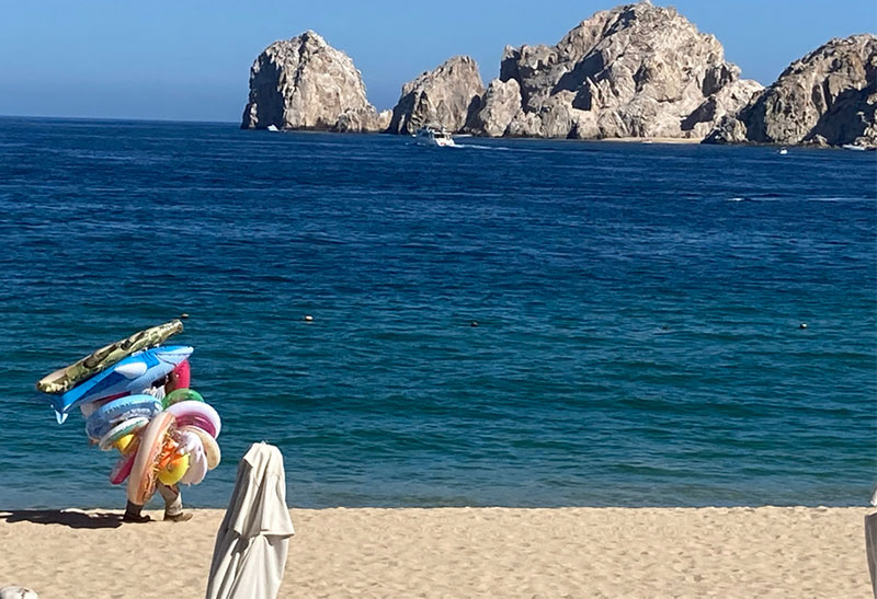 A view of Médano Beach on a sunny day, showing a beach vendor carrying a tall stack of hats walking along the sand, with the famous Land's End rock formations rising from the bright blue ocean in the background.
