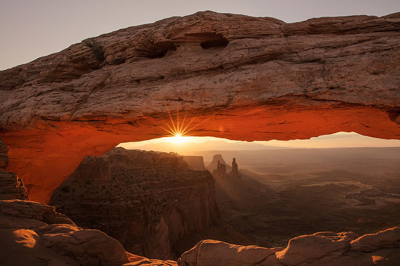 A breathtaking sunrise view through the stone opening of Mesa Arch in Canyonlands National Park, with the glowing orange underside of the arch framing the vast desert canyon and distant rock formations of Moab, Utah.