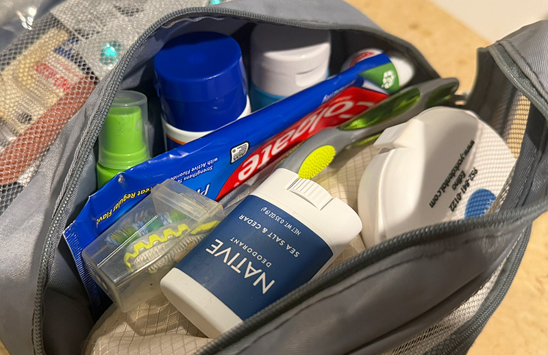 A high-angle, close-up shot of a small, clear plastic toiletry bag packed with travel-sized essentials. Inside the bag, a tube of Colgate toothpaste, a white Native deodorant stick, a small blue bottle, and other miscellaneous hygiene items are visible. The bag sits on a clean, white surface, illustrating a compact and organized approach to packing toiletries.