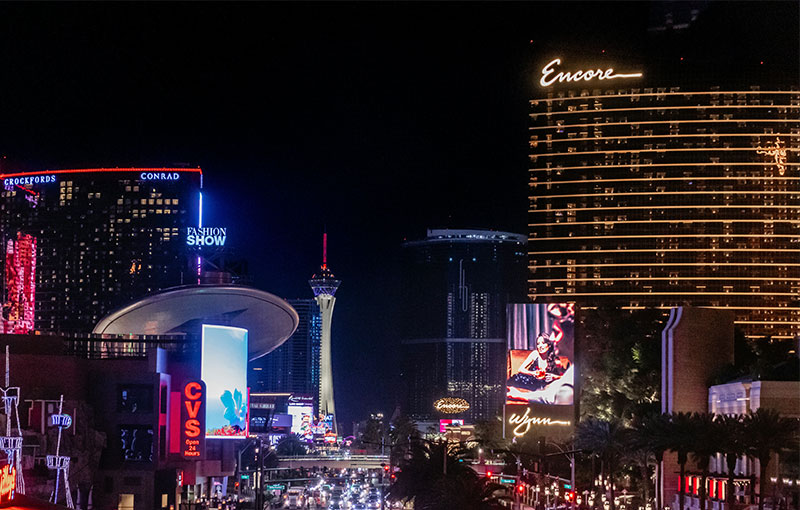 A vibrant nighttime view of the Las Vegas Strip, showcasing bright neon signs, bustling traffic, and prominent casino resorts like the Encore and the Stratosphere illuminated in the distance.