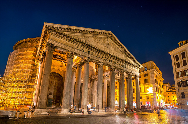 A wide-angle, low-angle shot of the Pantheon in Rome illuminated at night. The massive Corinthian columns of the portico are bathed in warm yellow light, casting deep shadows that emphasize their scale