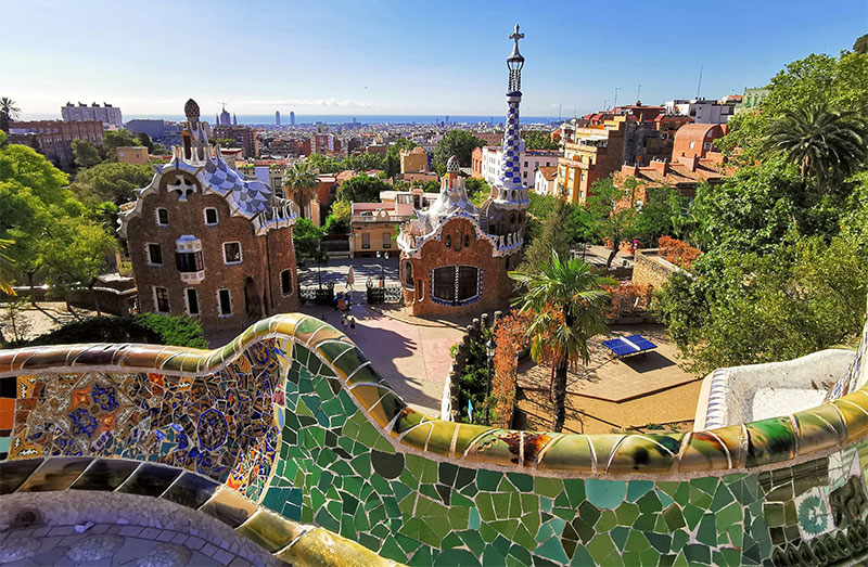 A panoramic view from the mosaic-covered terrace of Park Güell, showing the colorful gingerbread-style gatehouses and the city of Barcelona stretching toward the Mediterranean Sea.