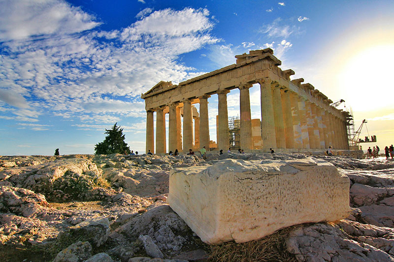 A wide scenic view of the ancient Acropolis of Athens at sunset, featuring the Parthenon temple standing prominently atop the rocky hill overlooking the city