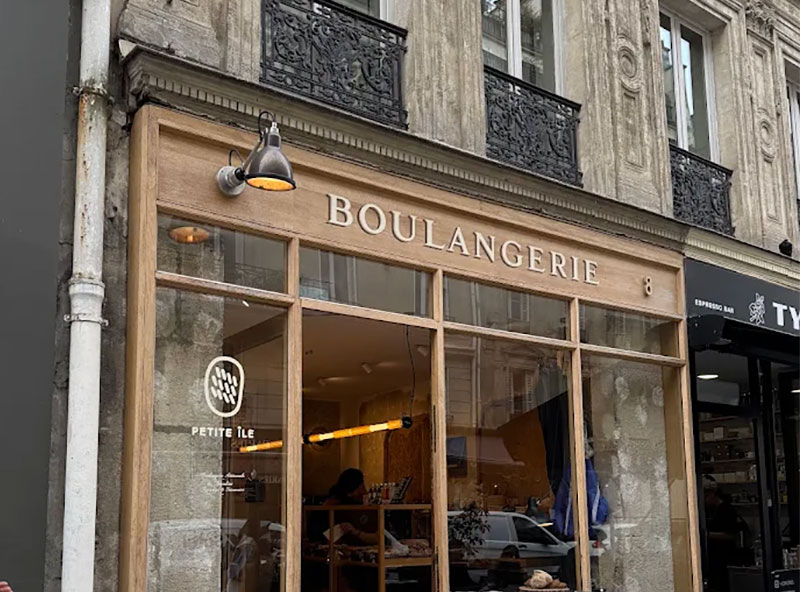 The storefront of Petite Ile boulangerie in Le Marais, featuring a light wood facade, large glass windows revealing the warm interior, and "BOULANGERIE" written in elegant lettering above the entrance
