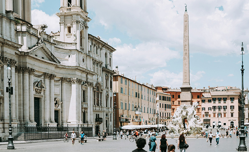 A wide-angle, eye-level shot of Piazza Navona in Rome during the blue hour. In the center, the Fountain of the Four Rivers (Fontana dei Quattro Fiumi) is illuminated, its carved marble figures and central Egyptian obelisk standing out against the deepening sk
