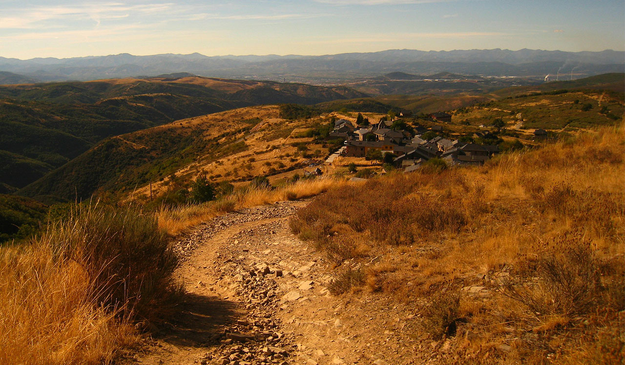 A winding dirt path through a grassy, mountainous landscape under a hazy sky during sunset or sunrise.