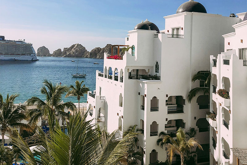 A view from a balcony showing a white Mediterranean-style resort building surrounded by palm trees, overlooking the calm ocean and the distant rugged rock formations of Land's End in Cabo San Lucas.