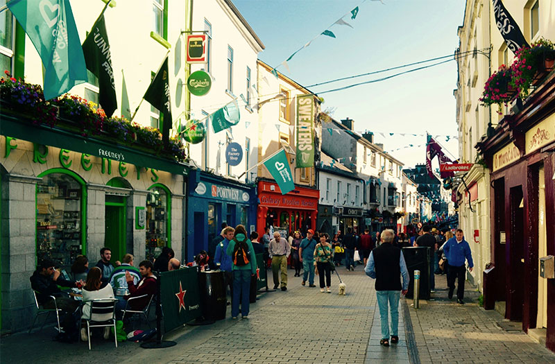 A lively view of Quay Street in Galway’s Latin Quarter, showing the colorful, brightly painted storefronts of traditional pubs and shops under a clear sky, with pedestrians walking along the historic cobblestone street.