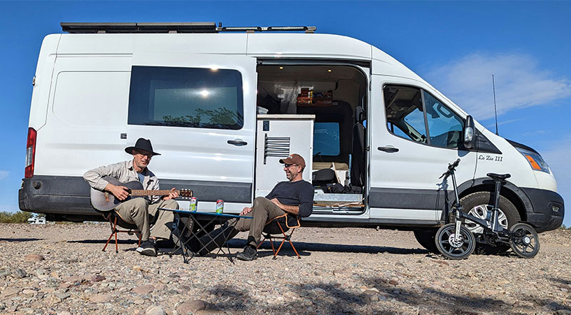 A traveler sits on a bench beside a white camper van with its side door open, parked in the open desert landscape of Quartzsite, Arizona, under a clear sky.