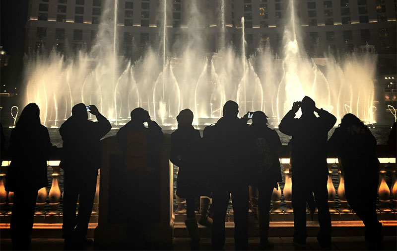A nighttime silhouette of a crowd of people watching the Bellagio Fountains, with high plumes of water illuminated against the dark sky and the glowing lights of the Strip in the background.