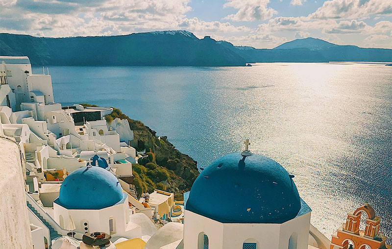 Alt Text: A classic view of Santorini, Greece, featuring a white-washed building with a bright blue dome in the foreground overlooking the deep blue Aegean Sea and the volcanic caldera under a soft, hazy sky.