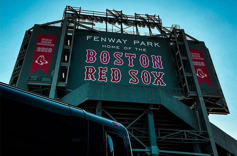 A wide view of the iconic green outfield and "Green Monster" wall at Fenway Park in Boston during a daytime baseball game, with the red seats of the grandstand filled with fans and the city skyline visible in the distance.