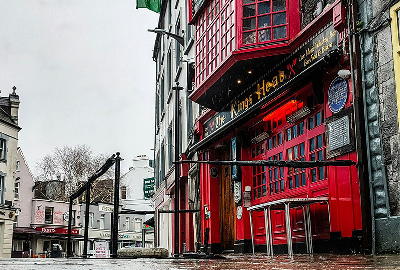 A vibrant street view of The King's Head pub in Galway, showcasing its bright red and yellow facade with traditional black signage, hanging flower baskets, and people walking by on the historic cobblestone street.