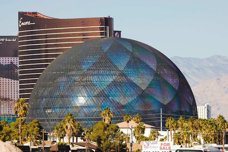 A wide exterior view of the Sphere in Las Vegas from Sands Avenue, featuring its massive LED exosphere displaying a glowing blue geometric pattern at dusk.