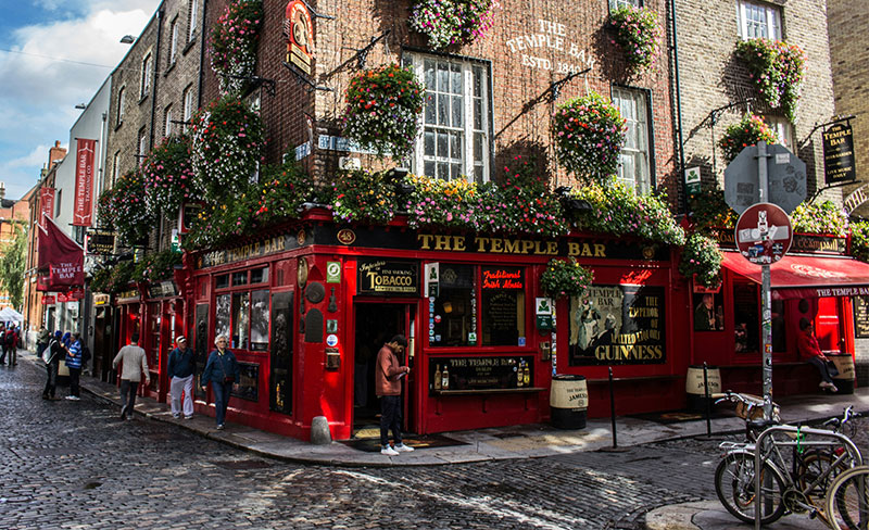 The iconic bright red exterior of The Temple Bar pub in Dublin, adorned with lush hanging flower baskets, gold lettering, and traditional black lanterns under a clear sky.