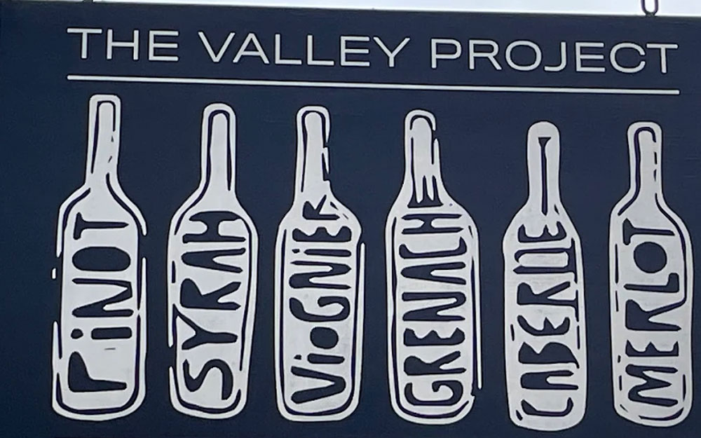 A large artistic mural on a chalkboard wall inside a wine tasting room, featuring five detailed wine bottle illustrations labeled with different grape varieties: Pinot, Syrah, Viognier, Grenache, Cabernet, and Merlot.