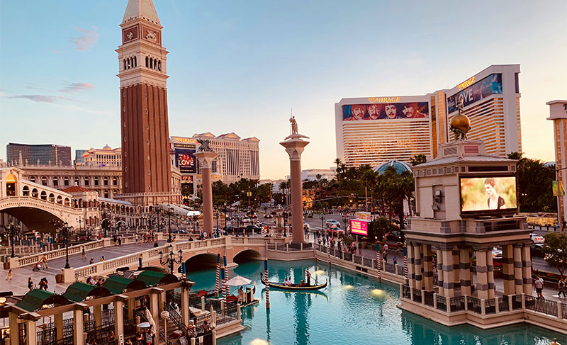 A wide daytime shot of The Venetian Resort in Las Vegas, showing the iconic Rialto Bridge replica, the Campanile tower, and the turquoise outdoor canals with gondolas.