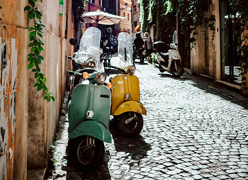 A vibrant, eye-level shot of a classic pastel-blue Vespa scooter parked on a narrow, sun-drenched Roman street. The scooter’s chrome details and tan leather seat gleam under the Mediterranean su