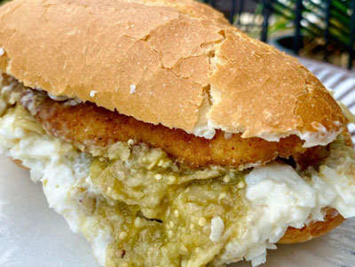 A close-up of a Torta de Chilaquil, featuring a crusty bolillo roll stuffed with green salsa-drenched chilaquiles, a breaded chicken cutlet (milanesa), and topped with a heavy drizzle of crema and crumbled queso fresco