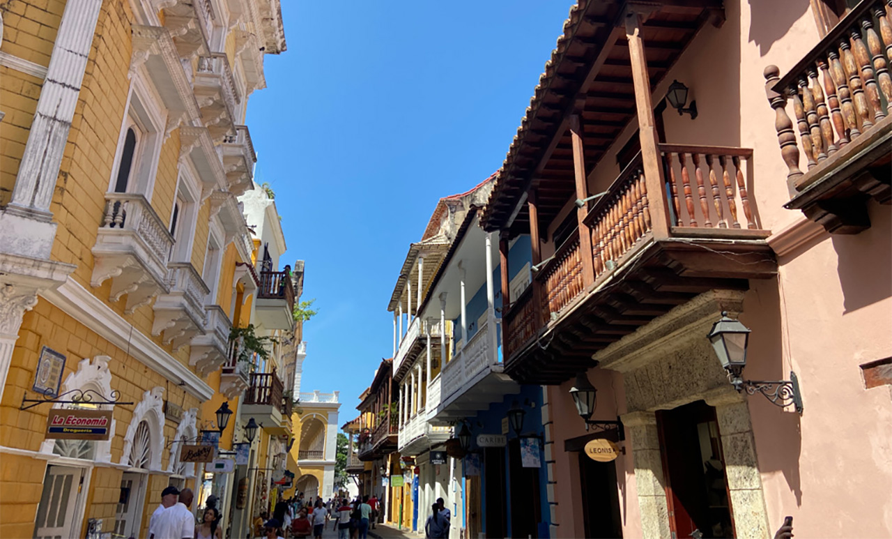 A narrow, sunlit street in the Walled City of Cartagena, Colombia, featuring colorful colonial buildings with traditional wooden balconies and vibrant bougainvillea flowers.