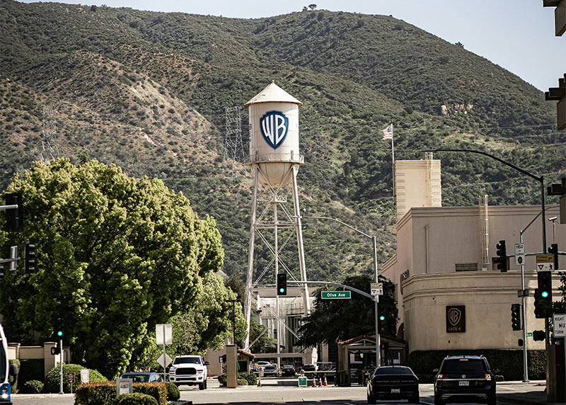The iconic white Warner Bros. Water Tower with the WB shield logo standing at the entrance of the studio backlot against a clear blue sky.