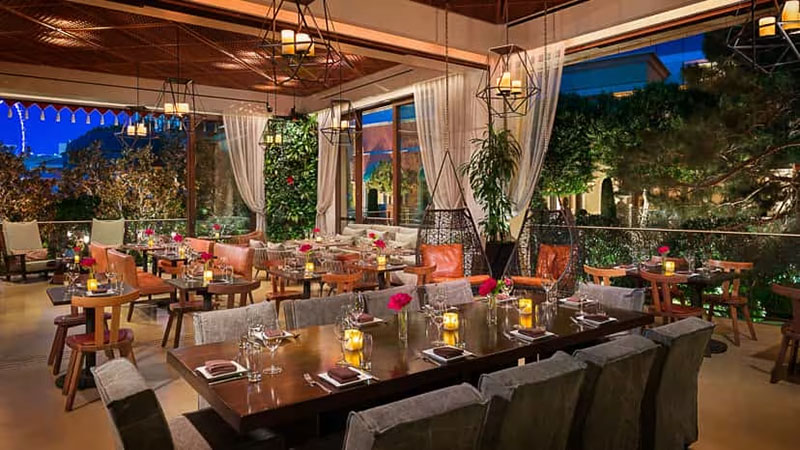 An elegant outdoor dining patio at night with a view of the Bellagio Fountains, featuring set tables under a dark wooden pergola with hanging lights.