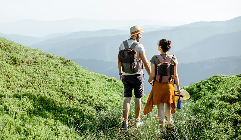 A man and woman standing on a grassy hilltop with backpacks, looking out at a scenic coastal view with blue water and distant cliffs under a clear sky.