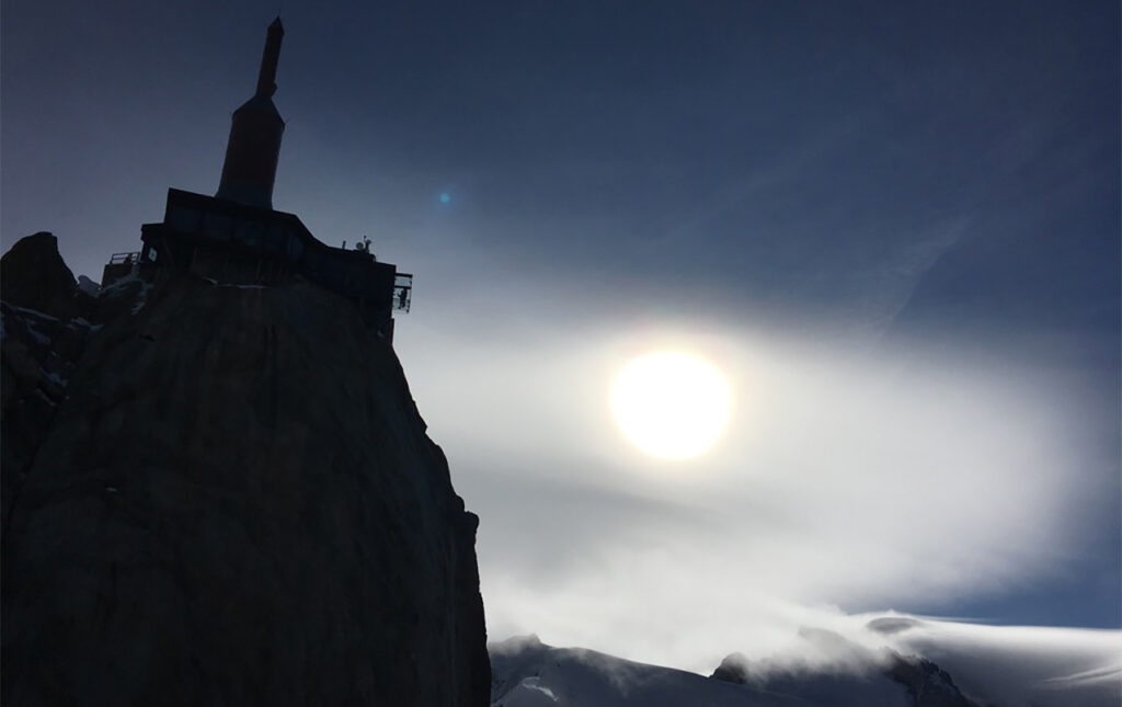 The silhouette of the Aiguille du Midi summit structure and its transmission tower against a bright sun and clouds.