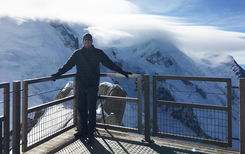 A man standing on a metal viewing platform at the Aiguille du Midi with a massive snow covered mountain peak behind him.