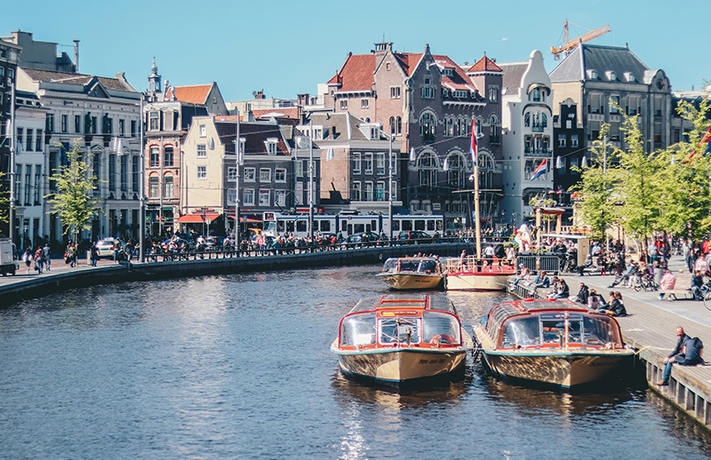 A row of historic, narrow gabled houses in various shades of grey and brick lining a calm Amsterdam canal, with several traditional wooden canal boats and small motorboats moored along the water’s edge under a clear sky.