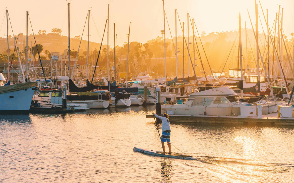 Paddleboarder navigating past anchored sailboats at the Santa Barbara Harbor during golden hour.