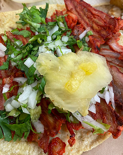 Close up of an authentic al pastor street taco topped with diced white onions, fresh cilantro, and a slice of pineapple on a yellow corn tortilla.
