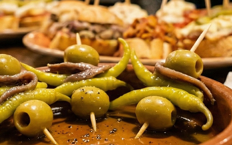 Traditional Basque Gilda pintxos with olives, guindilla peppers, and anchovies on toothpicks, served in a terracotta dish with olive oil on a wooden bar top.