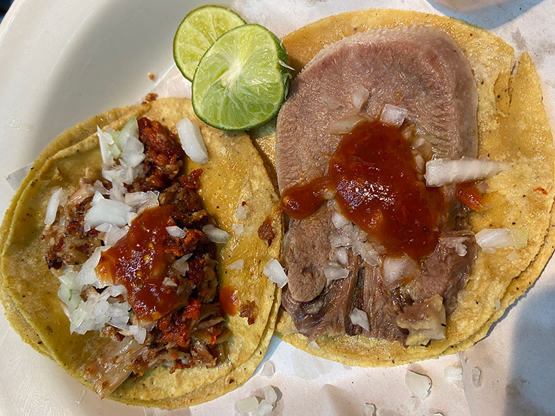 Two street tacos on a paper-lined plate, one featuring a thick slice of lengua topped with red salsa and onions