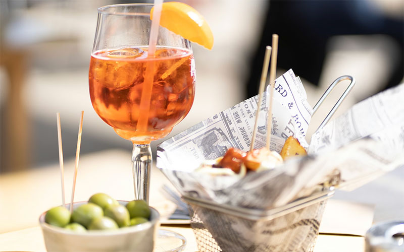 An Aperol Spritz cocktail with an orange slice alongside a basket of tapas and a small bowl of green olives on an outdoor cafe table.