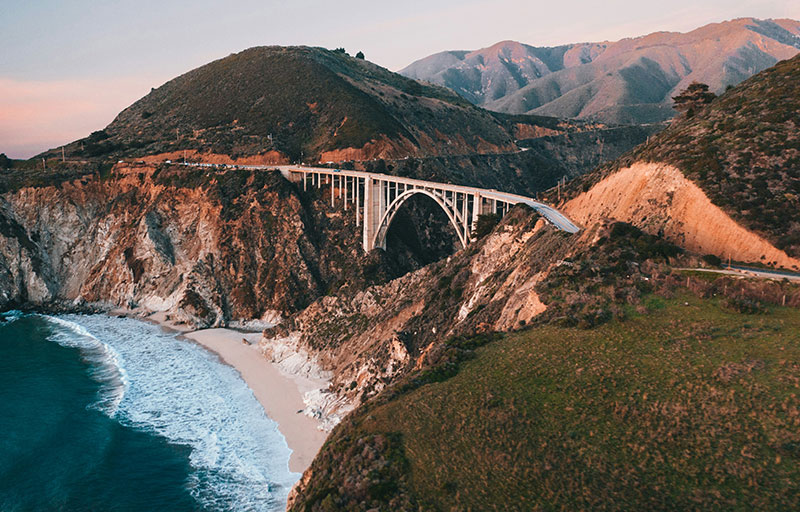 Aerial view of the historic Bixby Creek Bridge curving along the steep, rugged cliffs of the Big Sur coastline at sunset.