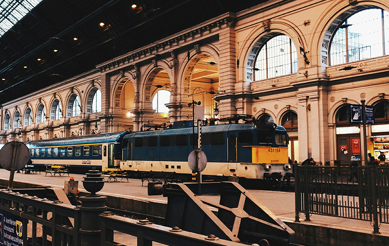 The interior of a grand, historic European train station in Budapest, featuring a high arched glass and steel ceiling, classic architecture, and a blue and yellow passenger train pulled up to a stone platform.