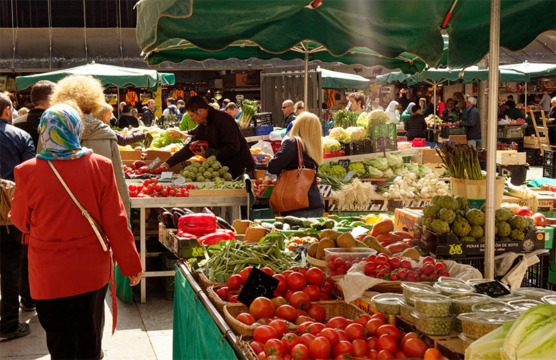 A busy outdoor European market with crowds of people browsing stalls overflowing with fresh red tomatoes, artichokes, and green vegetables under green awnings.