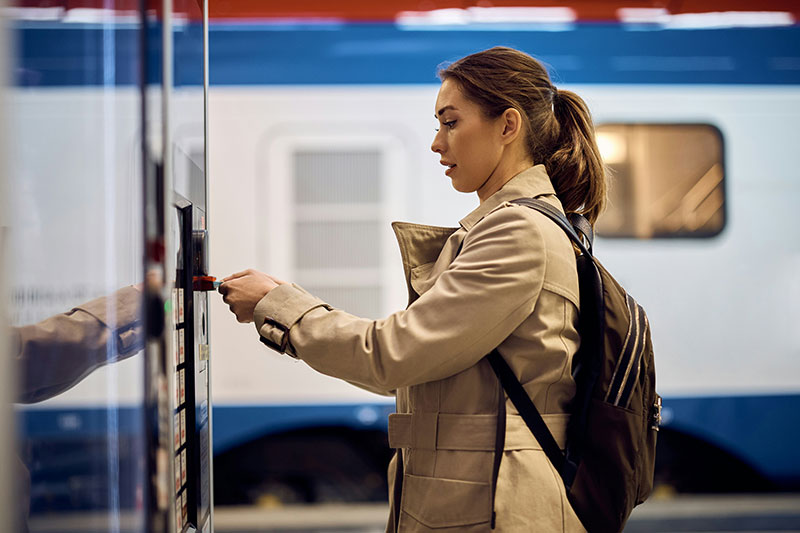 A woman in a tan trench coat standing on a train platform, looking up at a digital arrival and departure board with a focused and calm expression.