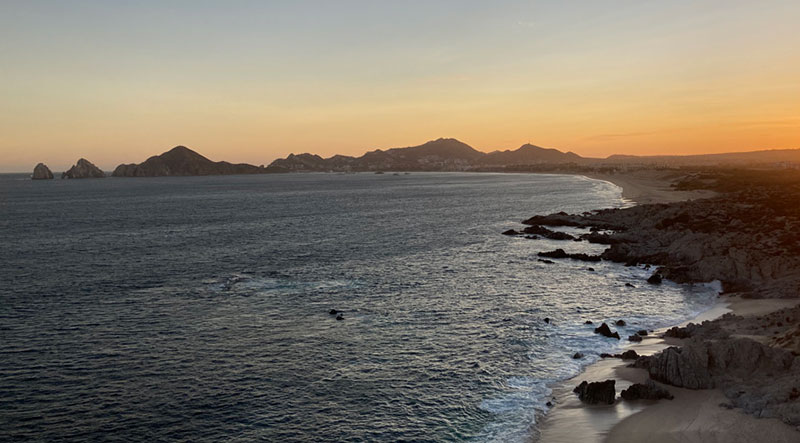 A vibrant orange and yellow sunset over the calm ocean, highlighting the rugged silhouettes of the coastline's rock formations in Cabo San Lucas.