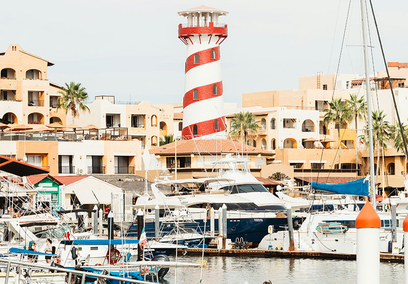 A view of a marina or harbor in Cabo San Lucas, Mexico, with several white boats docked. A distinctive red and white striped lighthouse stands prominently on the right. In the background are multi-story, white Spanish-colonial style buildings and palm trees, set against a bright blue sky with a few clouds.