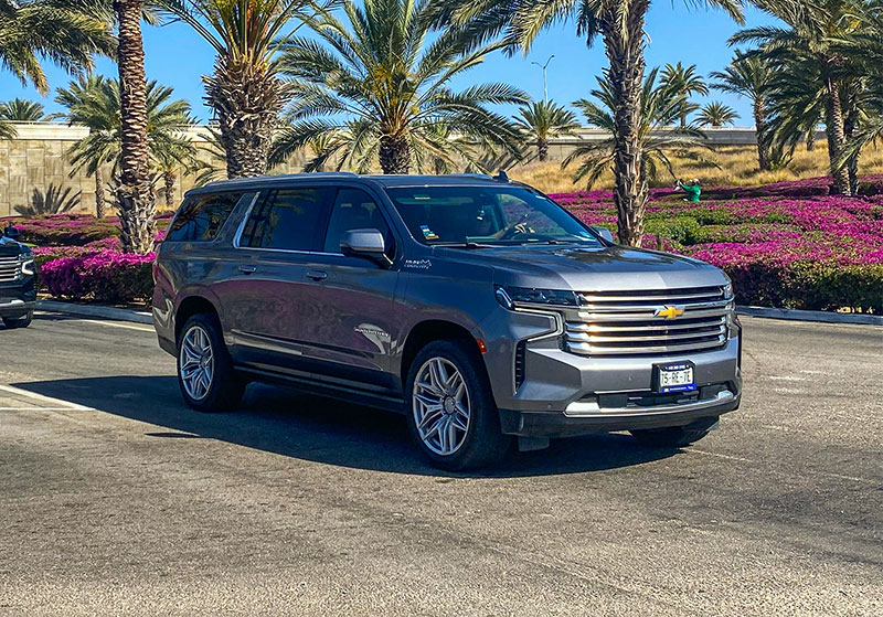A dark luxury SUV, commonly used for private airport transfers, parked on a sunny street lined with palm trees and resort buildings in Cabo San Lucas.