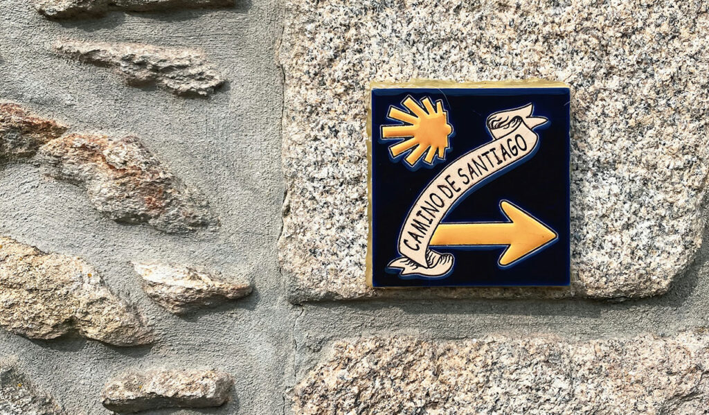 A blue ceramic tile embedded in a stone wall featuring the iconic yellow arrow and scallop shell pointing the way on the Camino de Santiago.