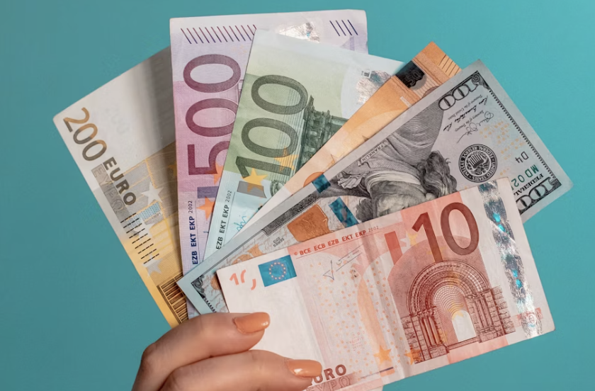 A hand holding a variety of colorful international banknotes, including Euro and British Pound notes, with a passport visible in the background.