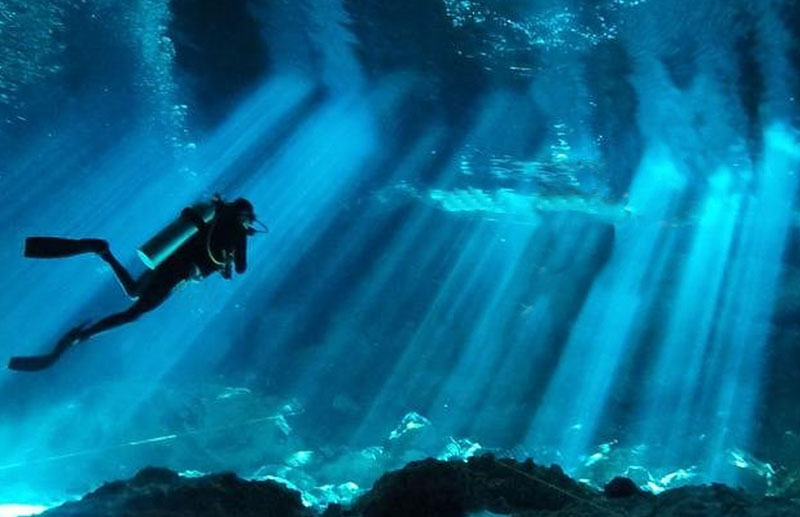 A scuba diver swimming through a Riviera Maya cenote with dramatic sunbeams, crystal-clear water, and limestone cave formations