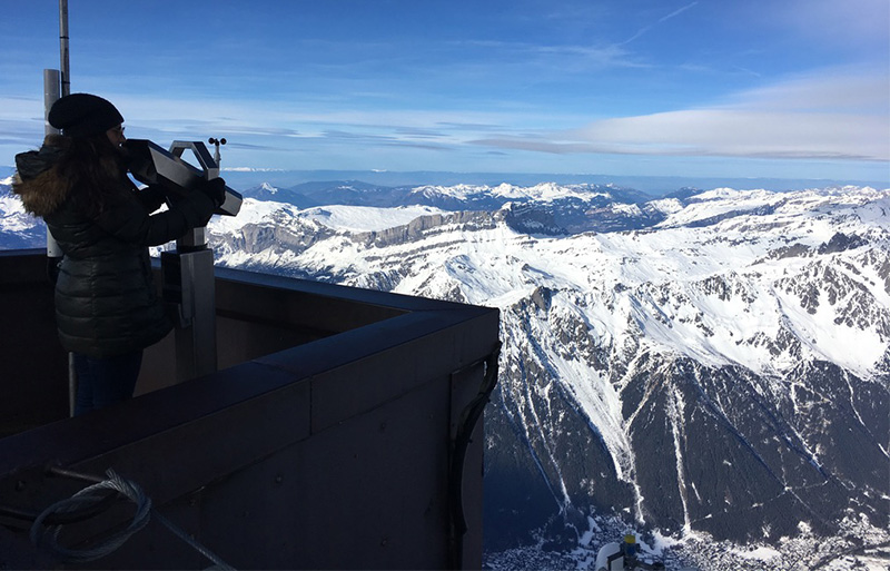 A woman looking through binoculars on a high altitude viewing platform overlooking snow capped Alpine peaks.