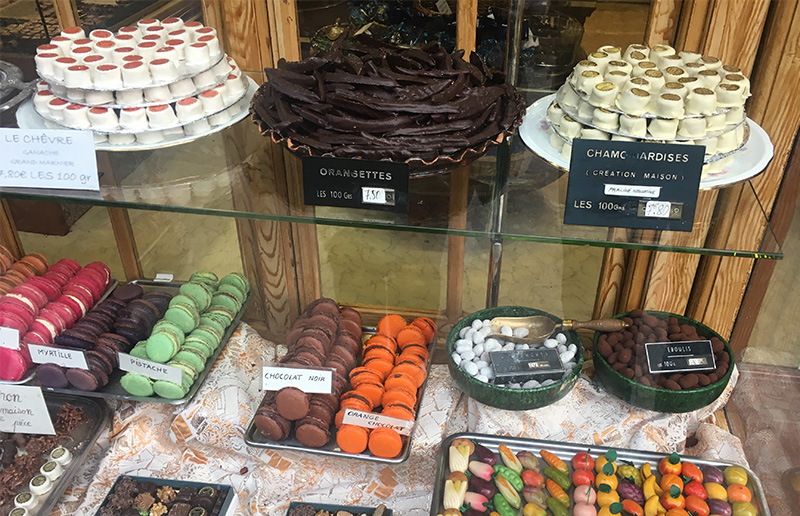 A window display at a French bakery featuring colorful macarons, chocolates, and marzipan shaped like fruit.