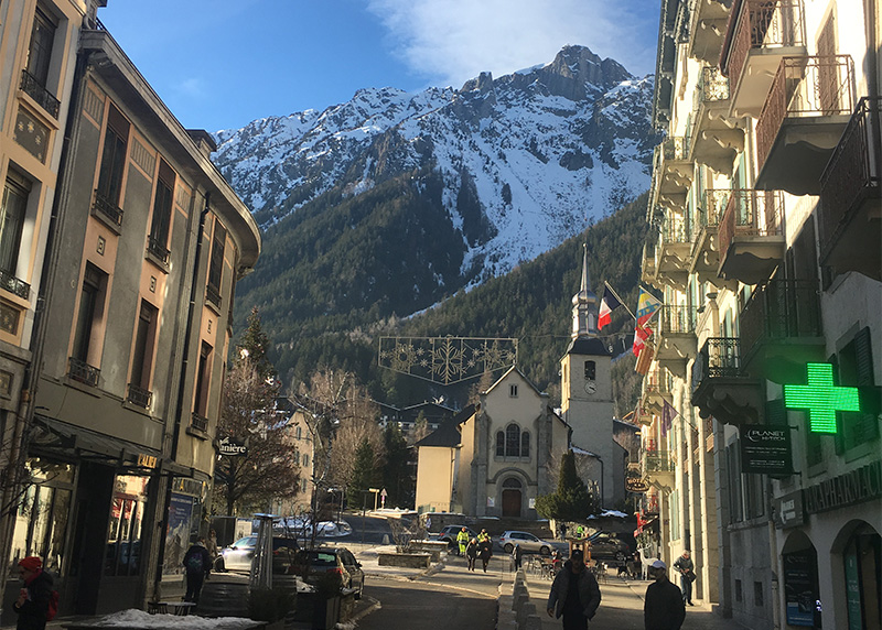 A street view of the Chamonix town center leading to the Eglise Saint-Michel with snow covered mountains in the background.