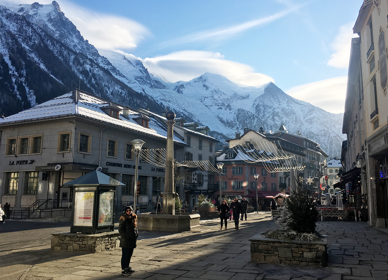 A woman standing in a sunny, cobbled square in the center of Chamonix, with historic buildings and the massive snow capped peaks of Mont Blanc towering in the background.