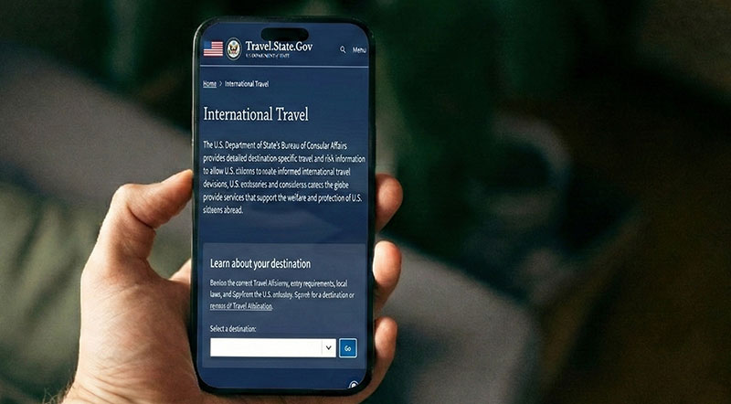 A person holding an iPhone displaying the U.S. Department of State's Travel.State.Gov International Travel website, used for checking destination-specific travel advisories.